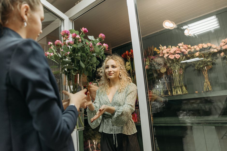A young woman with curly blonde hair, wearing a light-colored patterned blouse, stands inside a flower shop at Clerkenwell Florist. She is receiving a bouquet of pink roses with green foliage, wrapped