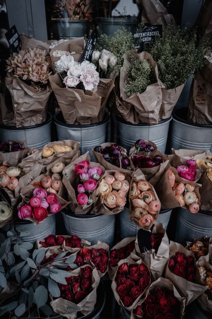 A collection of freshly cut flowers arranged in metal buckets, including tightly packed roses in shades of pink, peach, red, and cream, with some dark burgundy blooms. The bouquets are wrapped in brow
