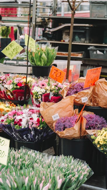 A market stall displaying a variety of fresh flower bouquets and individual blooms. Prominent are pastel-colored tulips with pink and white petals, arranged in black buckets on the lower part of the d