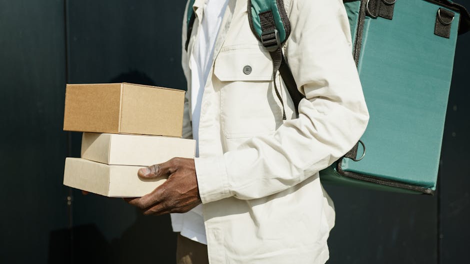 A person wearing a white jacket is holding three stacked rectangular cardboard boxes, with the top box being brown and the lower two white, in front of a dark background. The person's hand is visible,