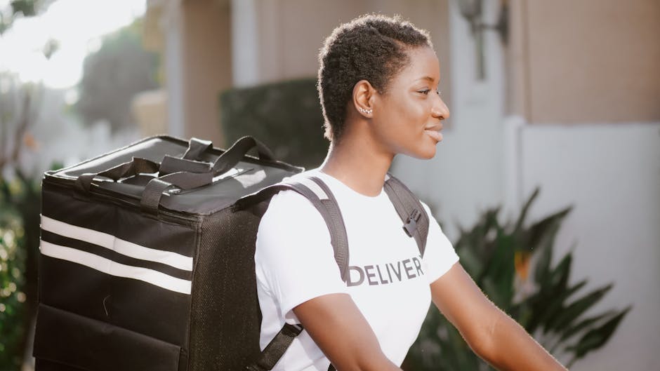 A female flower delivery courier with short, curly hair and a light brown complexion, smiling while riding a bicycle outdoors during daylight. She is wearing a white T-shirt with the word 'DELIVER' vi