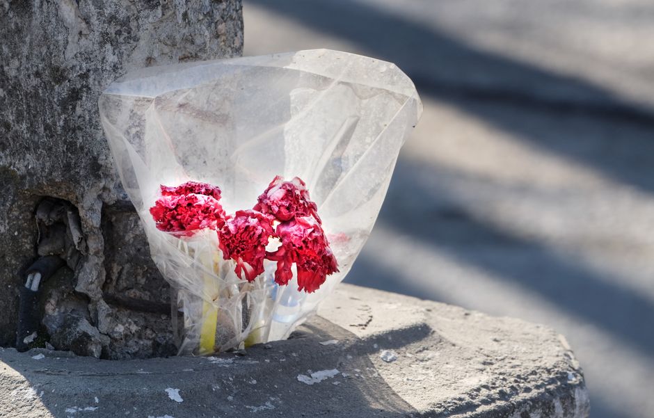 A small bouquet of pink carnations wrapped in clear cellophane, resting against a weathered stone wall on a concrete surface. The flowers appear slightly wilted with delicate, ruffled petals and visib